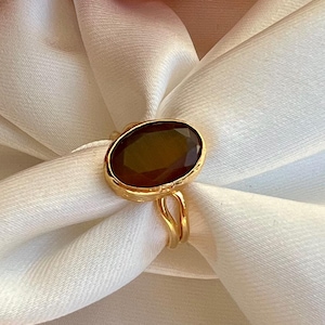May include: A gold ring with a large, oval, dark brown gemstone. The ring is set on a white fabric, with peach-colored flowers in the background. The ring's band is thin and delicate.