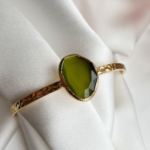 May include: A gold-coloured bracelet featuring a textured band and an olive-green gemstone. The gemstone is oval and set in a gold bezel. The bracelet is displayed on white, silky fabric, with flowers in the background.