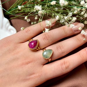May include: Two gold rings with teardrop-shaped gemstones, one ruby red and the other pale green, are displayed on a hand. The rings are set against a backdrop of white flowers and a brown surface, showcasing the jewelry's design.