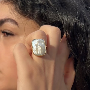 Puede incluir: Un anillo dorado con una gran perla barroca rectangular de color blanco iridiscente engastada en un bisel dorado liso. La perla tiene una textura superficial orgánica e irregular con un acabado brillante, y el aro es fino y de color dorado.