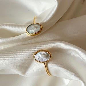 May include: Two gold-colored rings with oval, white, pearl-like stones. The stones are set in a gold bezel. The rings are displayed on a white, silky fabric, with dried lavender sprigs in the background. Jewelry.