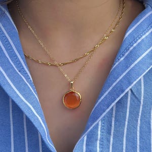 May include: A gold chain necklace with a round orange pendant. The necklace is layered over a blue and white striped shirt.