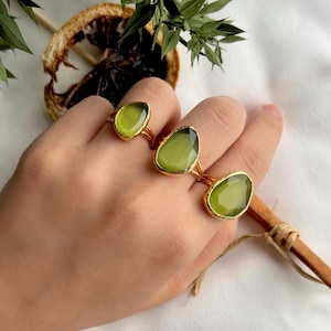 May include: Three gold rings with large, light green gemstone settings are displayed. The rings have a simple, elegant design, with the gemstones in oval and irregular shapes. The rings are on a hand, with a white background and decorative elements.