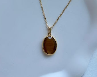 May include: A gold-colored necklace with an oval, brown gemstone pendant. The pendant is set in a gold frame and hangs from a delicate chain. A red ladybug figurine with black spots is in the background.