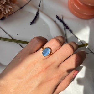 May include: A gold ring with a light blue oval gemstone. The ring is on a hand, with a soft background of flowers and other decorative items. The ring's design is simple and elegant.