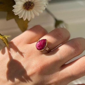 Op de afbeelding: Een gouden ring met een druppelvormige roze edelsteen. De ring zit aan een vinger.