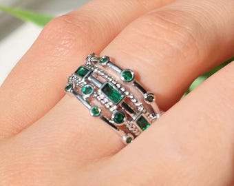 May include: A silver ring with multiple bands, adorned with emerald-green gemstones. The ring features a mix of round and rectangular stones, creating a layered and textured design. The ring is on a person's finger, with green nail polish visible in the background.