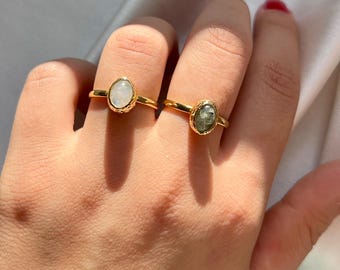 Puede incluir: Dos anillos dorados con piedras preciosas ovaladas. Un anillo presenta una piedra blanca, el otro una piedra verde grisácea. Los anillos tienen una banda dorada texturizada. Los anillos se muestran en una mano.