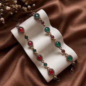 May include: A gold-toned metal chain bracelet featuring alternating teardrop-shaped red and green stones set in ornate, textured bezels. The bracelet includes a lobster claw clasp and an adjustable extender chain with small circular links.