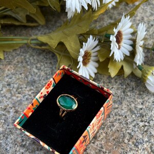 May include: A gold ring with a large oval green gemstone set in a gold setting. The ring is in a small box with a colorful patterned design.