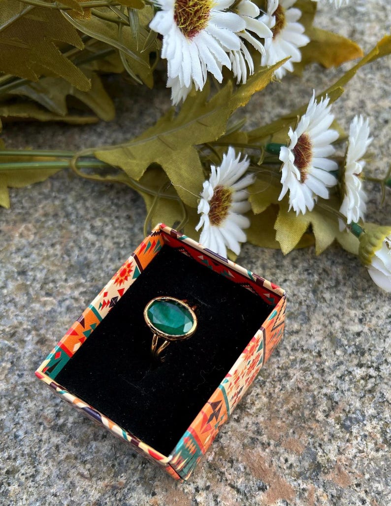 May include: A gold ring with a large oval green gemstone set in a gold setting. The ring is in a small box with a colorful patterned design.