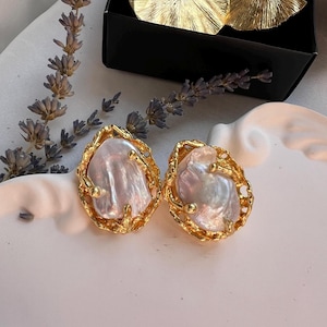 May include: A pair of gold-coloured earrings featuring large, irregularly shaped, pearlescent stones. The earrings have a textured gold border with small gold accents. The earrings are displayed on a white surface with lavender sprigs and a black box in the background.