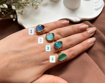 May include: A hand displays four gold rings with oval-shaped gemstones in blue and green hues. Two additional rings with similar designs are displayed on a white ceramic stand. The rings are set against a brown fabric background.