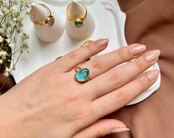 May include: A gold ring with a teardrop-shaped blue gemstone. The ring is on a finger of a hand with light skin tone.
