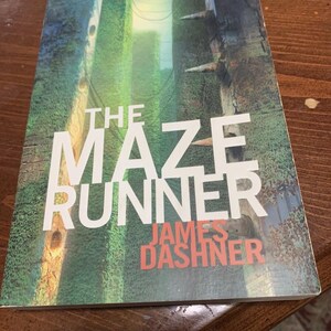 May include: A paperback book titled The Maze Runner by James Dashner. The cover features a green, textured, vertical maze-like structure with sharp, protruding spikes. The title is printed in large white letters, and the author's name is in red.