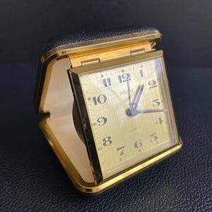 Op de afbeelding: Een vintage reiswekker met een goudkleurige vierkante wijzerplaat en zwarte cijfers. Op de wijzerplaat staan de woorden DELUXE en 2 JEWELS gedrukt. De wekker zit in een zwarte, gestructureerde opvouwbare etui met een goudkleurig metalen frame.