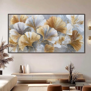 Peut inclure: Grande œuvre d'art encadrée représentant des feuilles de ginkgo stylisées dans des tons or, blanc et gris. L'œuvre est exposée au-dessus d'une console moderne dans un salon. La pièce comprend un canapé beige, des poufs bruns et un tapis aux tons neutres.