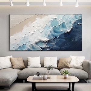 Puede incluir: Pintura abstracta de olas del océano rompiendo en una playa de arena. La pintura está en tonos de azul, blanco y beige. Se muestra en una pared sobre un sofá gris con almohadas.