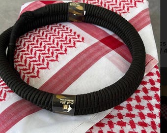 May include: A black rope-like headpiece, or agal, rests on a red and white checkered scarf. The agal has two gold-colored metal clasps with Arabic text. The scarf has a repeating geometric pattern in red and white.