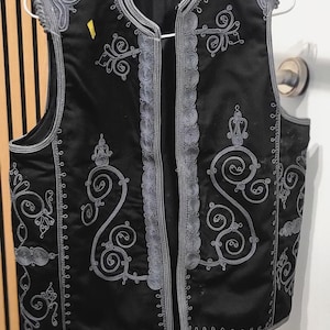 Può includere: Gilet nero con ricami grigi ornati. Il gilet presenta un'intricata lavorazione a volute e finiture decorative sui bordi e sul davanti. È senza maniche e appeso a un'appendiabiti su uno sfondo neutro.
