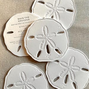 May include: White sand dollar-shaped wedding invitations and coasters. The invitations have the text "SAVE THE WEEKEND" and wedding details. The coasters are embossed with a sand dollar design.