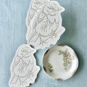 Op de afbeelding: Drie witte papieren uitsparingen van vogels, met gedetailleerde lijnkunst, zijn gerangschikt met een klein, rond bord in antieke stijl. Het bord heeft een bloemenpatroon en een gouden rand. De achtergrond is lichtblauw.