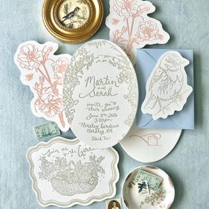 Op de afbeelding: Een verzameling trouwbriefpapier, waaronder uitnodigingen, RSVP-kaarten en postzegels. De uitnodigingen tonen bloemmotieven en de namen "Martin en Sarah". Een kleine ingelijste vogelkunst en een decoratief schaaltje zijn ook aanwezig. Het kleurenpalet is zacht en elegant.