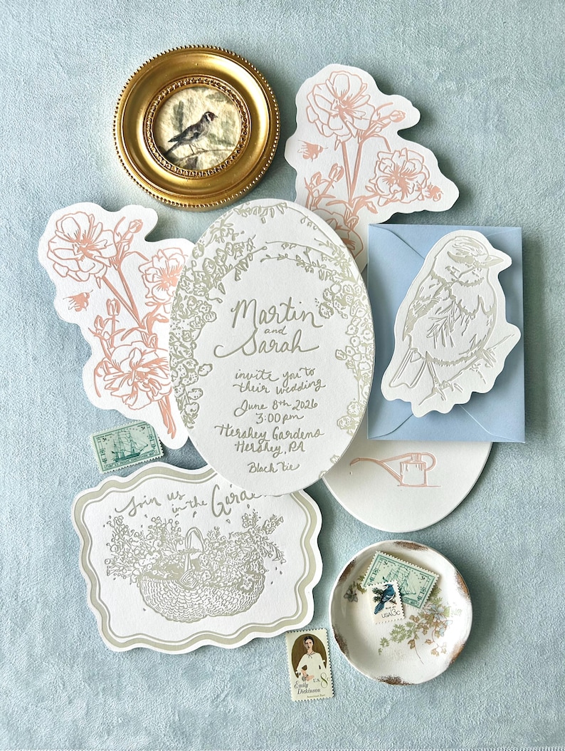 Op de afbeelding: Een verzameling trouwbriefpapier, waaronder uitnodigingen, RSVP-kaarten en postzegels. De uitnodigingen tonen bloemmotieven en de namen "Martin en Sarah". Een kleine ingelijste vogelkunst en een decoratief schaaltje zijn ook aanwezig. Het kleurenpalet is zacht en elegant.