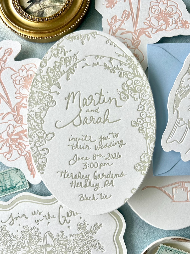 Op de afbeelding: Een trouwuitnodigingssuite met een ovale uitnodiging in het midden, met de namen "Martin en Sarah" en evenementdetails. De uitnodiging is omgeven door een bloemenontwerp. Andere briefpapierartikelen zijn inbegrepen.