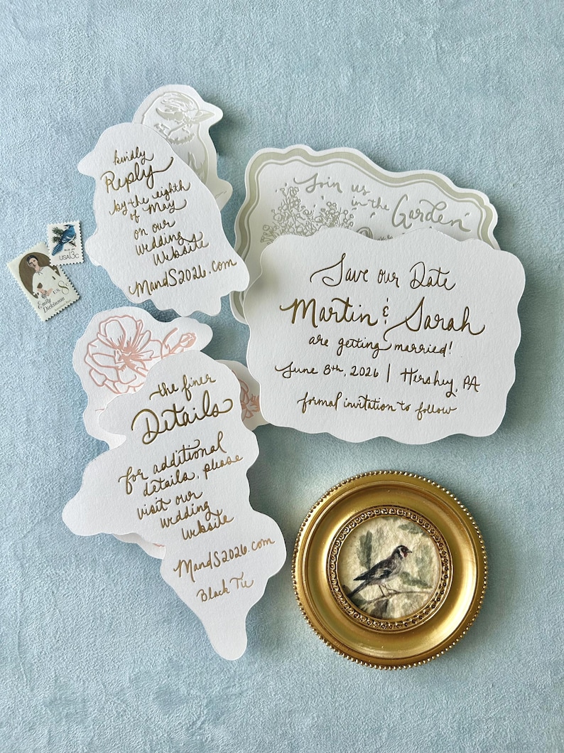 Op de afbeelding: Bruiloft uitnodigingsset met een goudkleurig thema. Bevat verschillende uniek gevormde kaarten met handgeschreven kalligrafie, een kleine ingelijste vogel schilderij en twee vintage postzegels. De kaarten bevatten de tekst "Save our Date" en "Join us in the Garden".