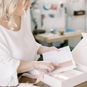 Op de afbeelding: Een persoon houdt een witte doos vast met een brochure en kleurstalen. De persoon draagt een witte blouse en gouden sieraden. Een roze lint ligt op de tafel. De achtergrond is een kamer met een houten tafel en een raam.