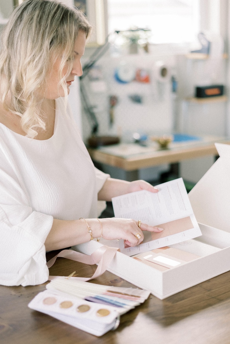 Op de afbeelding: Een persoon houdt een witte doos vast met een brochure en kleurstalen. De persoon draagt een witte blouse en gouden sieraden. Een roze lint ligt op de tafel. De achtergrond is een kamer met een houten tafel en een raam.