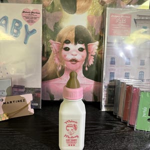 May include: A white plastic bottle with a pink cap and a brown nipple. The bottle has a label that says "Melanie Martinez Cry Baby Perfume Milk".