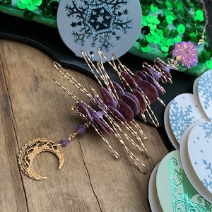 May include: A gold wire Christmas tree ornament with purple iridescent shell-like ornaments and a gold crescent moon charm. The ornament is hanging from a gold wire.
