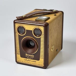 Peut inclure: Un appareil photo box vintage Kodak Brownie Six-20 Model F avec un corps beige et une façade marron foncé. L'avant présente deux viseurs circulaires, un objectif central et le texte Brownie Six-20 Camera Model F with flash contacts.
