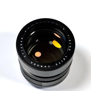 May include: A black camera lens with a circular glass element featuring orange and white reflections. The outer ring is inscribed with the text LEITZ CANADA and 1:2/90, along with a numerical scale ranging from 2 to 40 in white lettering.
