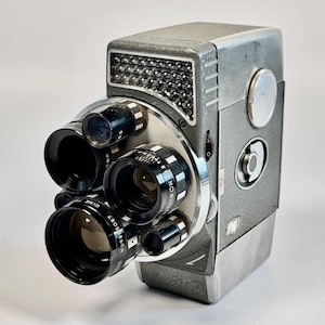 May include: A vintage silver and grey 8mm movie camera featuring a rotating turret with multiple black lenses. The body has a textured finish, a rectangular light meter window with a grid pattern, and various manual adjustment dials.