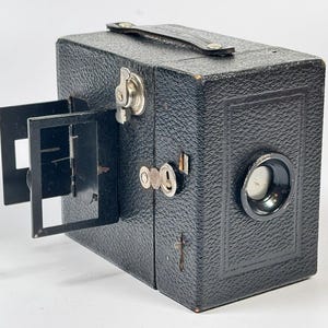 May include: A vintage black box camera with a textured leatherette finish, featuring a top carrying handle, a side-mounted metal viewfinder frame, a circular lens, and silver-toned metal hardware including a shutter release and latch.
