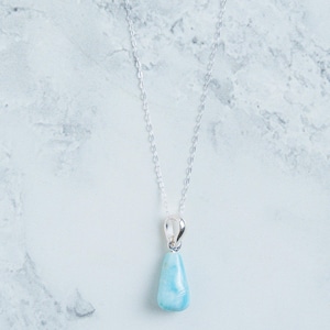 May include: A silver necklace with a teardrop-shaped, light blue pendant. The pendant has a marbled appearance, and the chain is delicate. The necklace is displayed against a white and gray marble background.