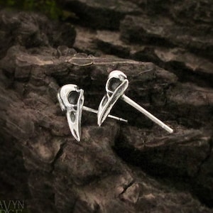 May include: A pair of silver earrings featuring small bird skull designs. The earrings are on a wooden surface.