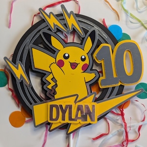 May include: A yellow Pikachu cake topper with a black and grey lightning bolt design. The topper also includes the name "DYLAN" and the number "10" in silver.