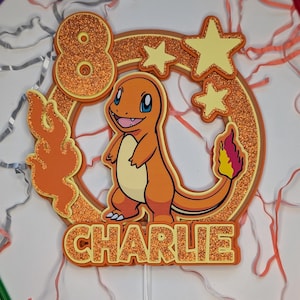 Puede incluir: Un adorno para pastel personalizado con un personaje de Charmander, el número 8 y el nombre "Charlie". El diseño incluye detalles de purpurina naranja, estrellas amarillas y un detalle de llama. El adorno está montado en un palito de plástico transparente.