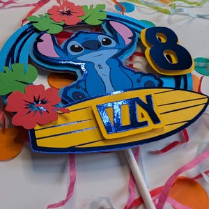 May include: Blue Stitch character sitting on a yellow surfboard with a blue "8th Day" banner. The character is surrounded by green leaves and red flowers.