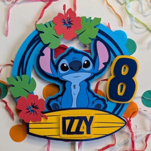 May include: Blue Stitch character cake topper with a surfboard, red hibiscus flowers, green leaves, and the name "Izzy" on the surfboard. The number 8 is on the side of the topper.