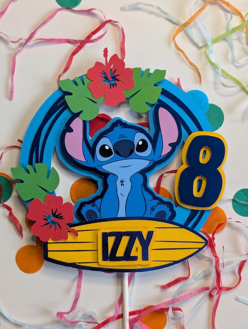 May include: Blue Stitch character cake topper with a surfboard, red hibiscus flowers, green leaves, and the name "Izzy" on the surfboard. The number 8 is on the side of the topper.
