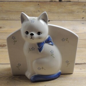 May include: A white ceramic napkin holder featuring a sculpted cat sitting in the center. The cat has a blue bow around its neck, a blue tail, and small blue floral patterns on its body and the arched back panel of the holder.