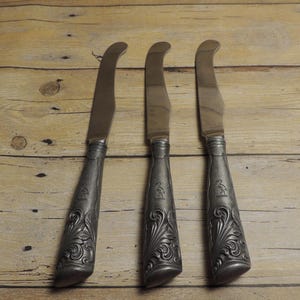 May include: Three silver-toned knives with curved blades and ornate, embossed metal handles featuring a floral pattern and a small bird emblem. The knives have a metallic finish and are arranged vertically, showcasing detailed craftsmanship on the handles.
