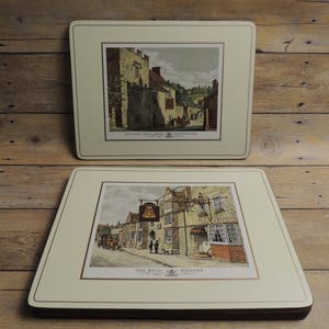 May include: Two rectangular placemats with cream-colored borders and central illustrations of historic English inns. The top placemat features a stone building labeled The Lord Crewe Arms, while the bottom placemat depicts The Bell at Stilton.