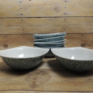 May include: A set of six ceramic bowls featuring a white interior and a speckled blue-grey exterior. The bowls have a distinct curved, wide-rimmed shape with pointed edges on opposite sides, creating a modern silhouette for serving food.