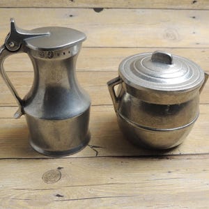 May include: A silver-toned metal creamer pitcher with a hinged lid and handle alongside a matching lidded sugar bowl. The pitcher features the text USA stamped near the rim. Both items have a smooth, matte finish and a rounded, functional design.